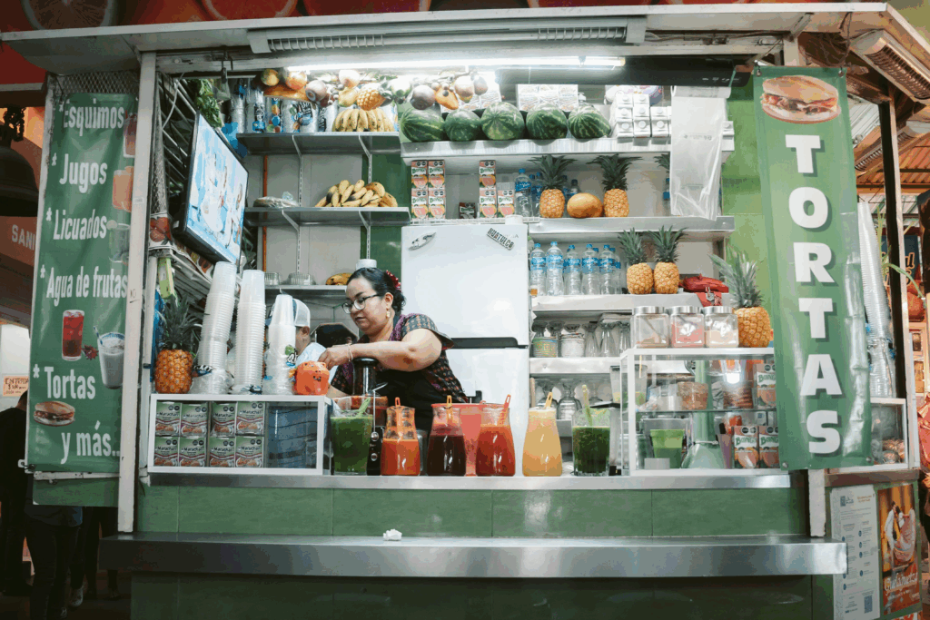 A woman prepares drinks at a vibrant juice and torta stand in Mexico City, with colorful jars of aguas frescas—red, green, and orange—lined up on the counter. Behind her, shelves overflow with fresh fruit like pineapples, bananas, and watermelons, creating a lively and inviting street food scene.