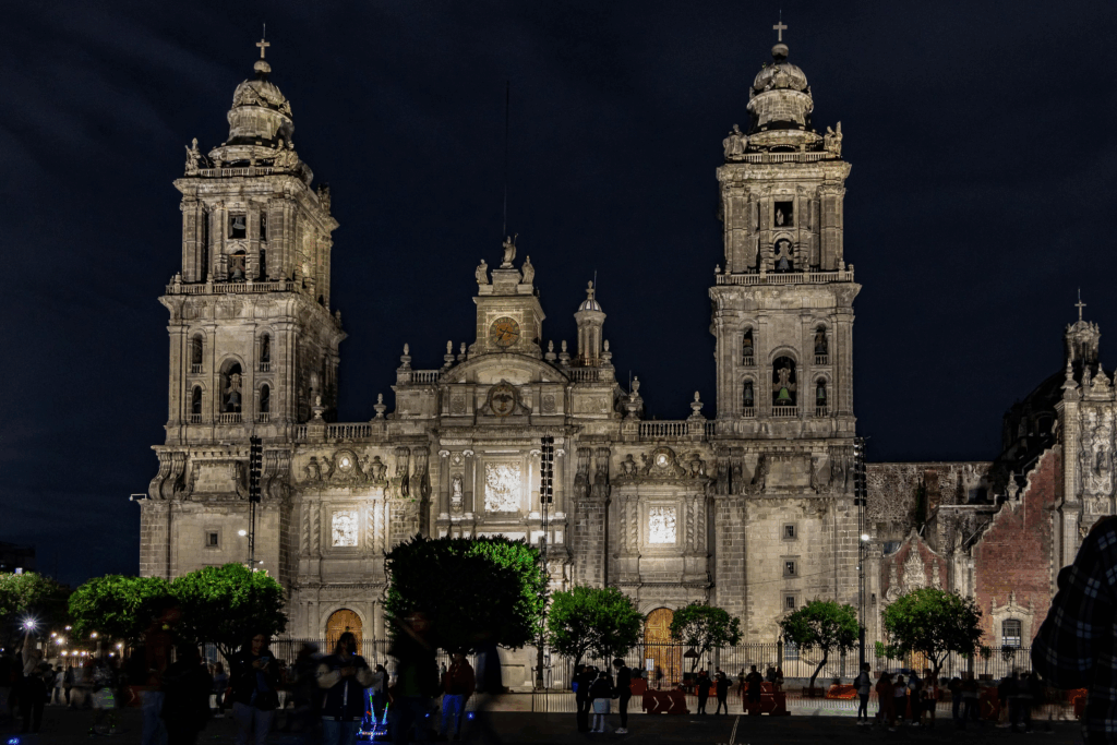 Night view of the Metropolitan Cathedral in Mexico City, dramatically lit against the dark sky. The Baroque and Neoclassical façade glows with golden light, highlighting intricate architectural details and soaring bell towers.