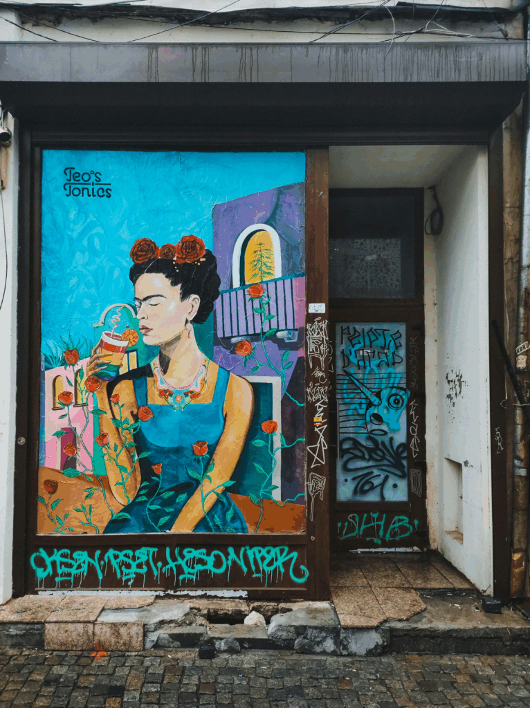 Street art mural of Frida Kahlo on a storefront in Mexico City. Set against a bright turquoise background, Frida is depicted in bold colors with flowers in her hair and a parrot on her shoulder. The piece is framed by a dark doorway and sidewalk, adding contrast to the vibrant artwork.