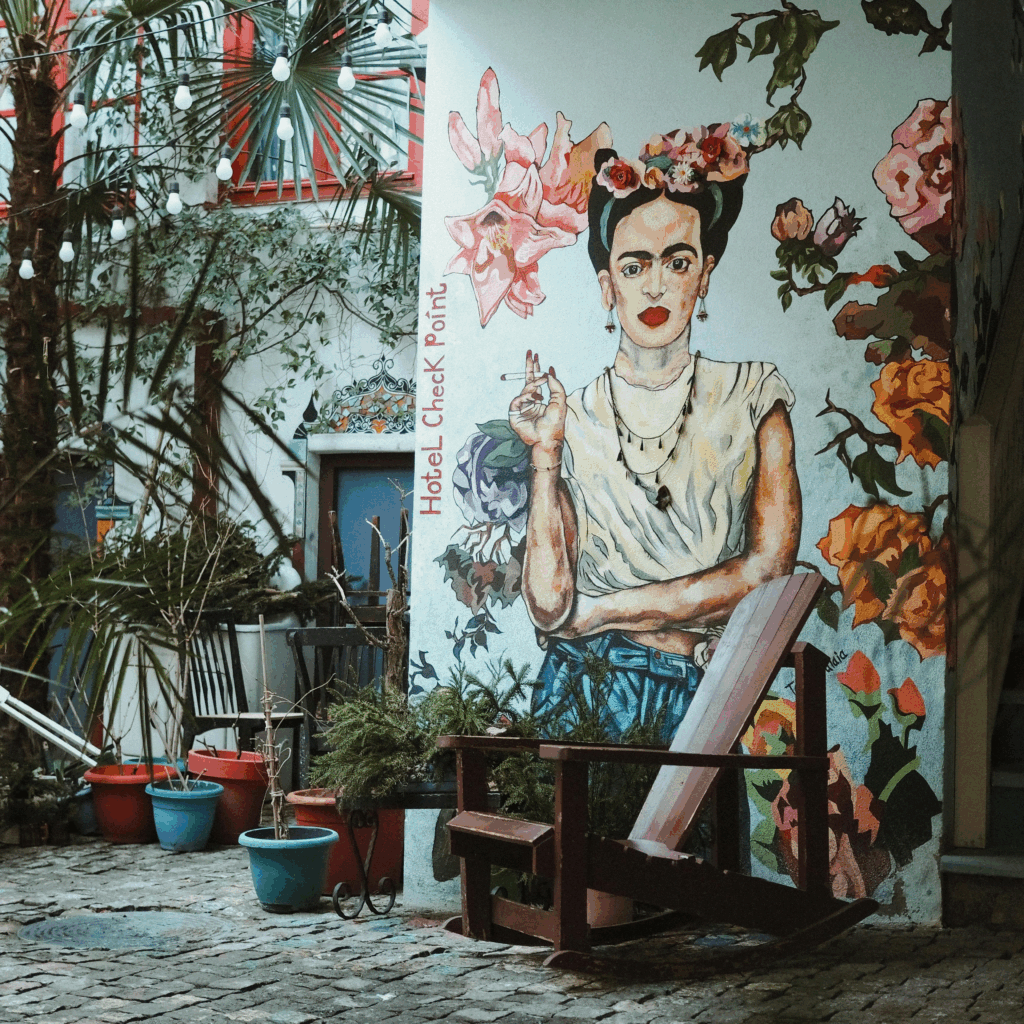Vivid street mural of Frida Kahlo in Mexico City, surrounded by bold floral elements and tropical greenery. The iconic artist is depicted seated with her signature unibrow, floral crown, and traditional Tehuana dress, blending into a lush, urban garden backdrop.