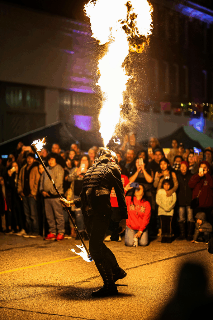 A fire performer in Mexico City breathes a dramatic burst of flame into the night sky, captivating a large crowd gathered around. The fiery plume glows against the dark street, while spectators watch in awe beneath colorful festival lights.