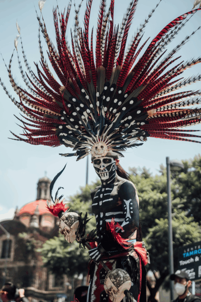 A Día de los Muertos performer in Mexico City wears elaborate face paint resembling a skull and a towering feathered headdress. Dressed in traditional black attire with bone-like designs, they hold a red flower, standing against a backdrop of colonial rooftops and greenery.