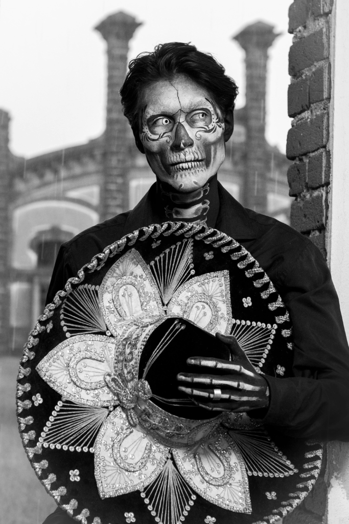 Black and white portrait of a Día de los Muertos performer in traditional mariachi attire, with skeletal face paint and a decorated sombrero held across their chest. The serious expression and monochrome palette evoke reverence and timeless cultural pride.
