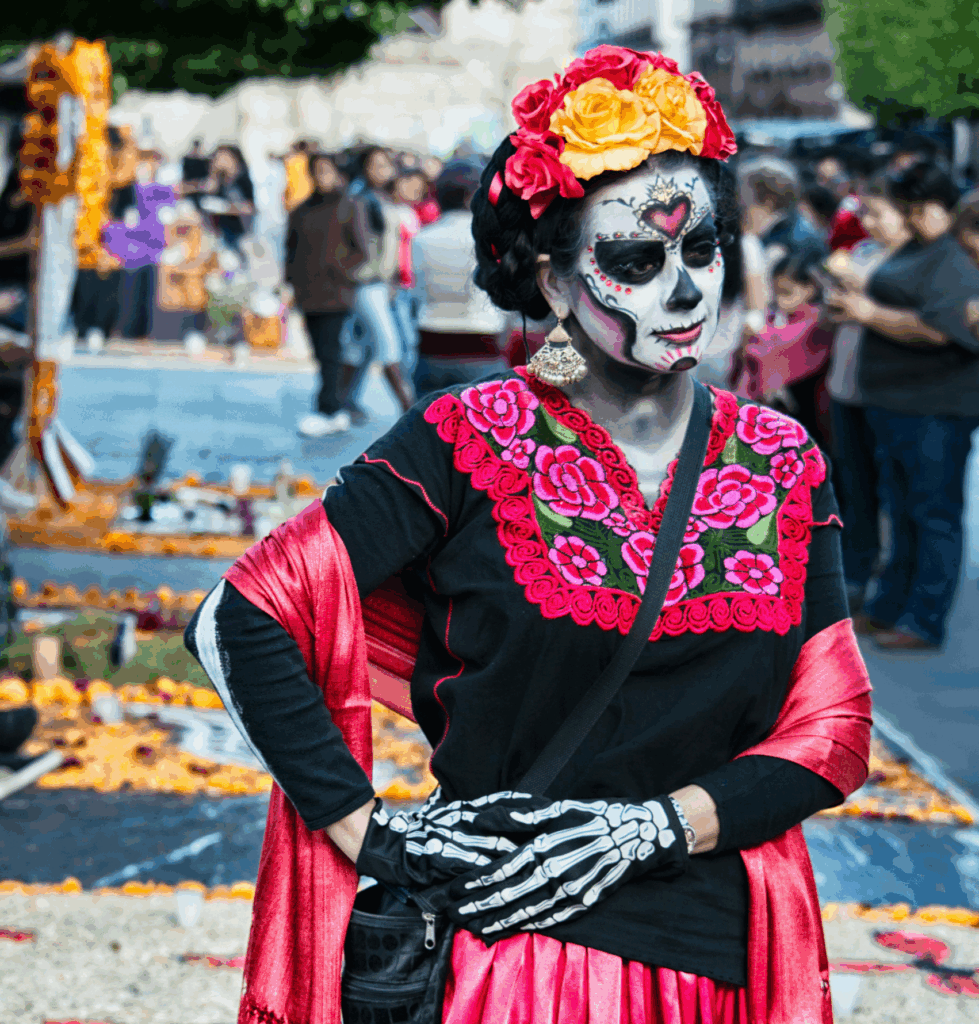 A woman dressed for Día de los Muertos wears a vibrant red and black embroidered dress, with detailed skeletal face paint and gloved hands posed like a calavera. She stands before a colorful altar filled with marigolds, candles, and offerings, honoring the tradition of remembering the dead.